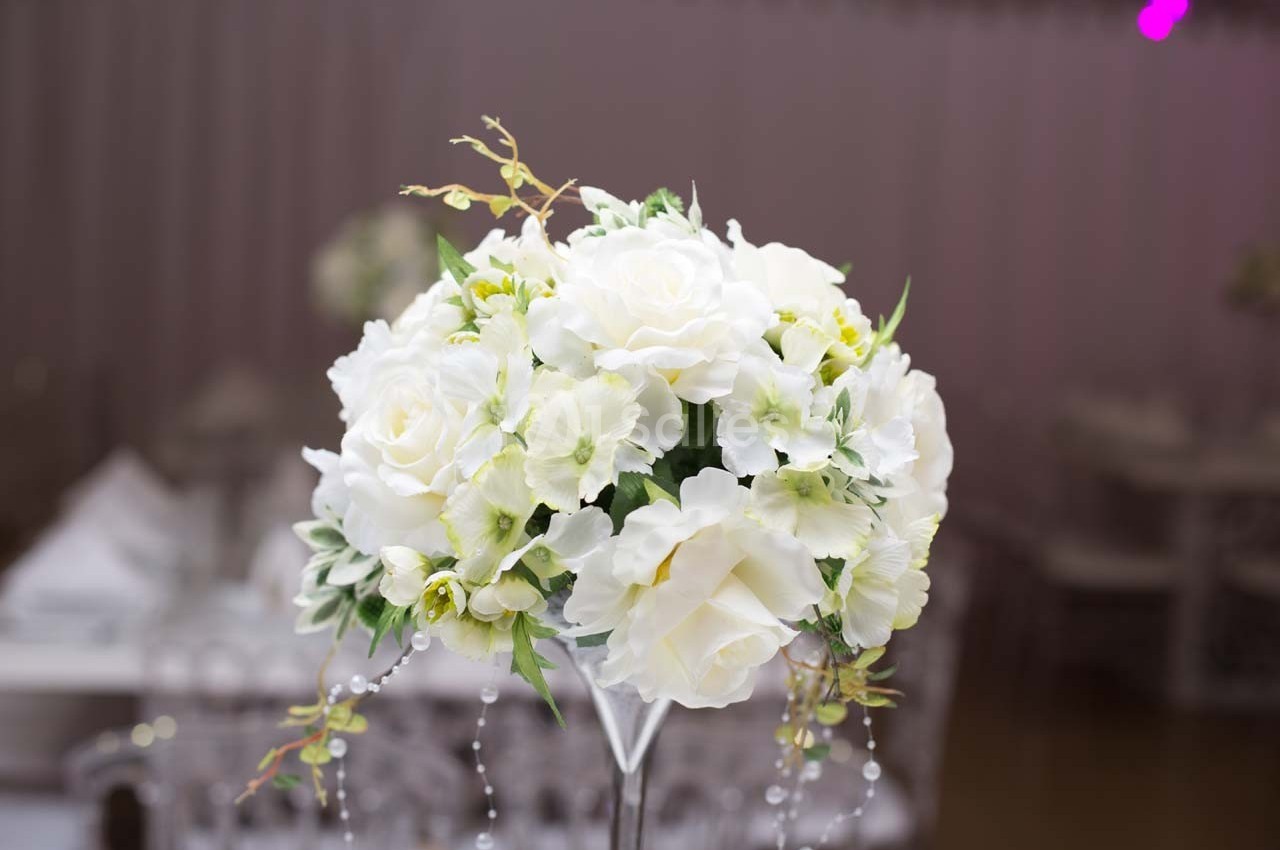Bouquet de fleurs blanches avec des perles décoratives, posé sur un support transparent dans un décor intérieur flou.