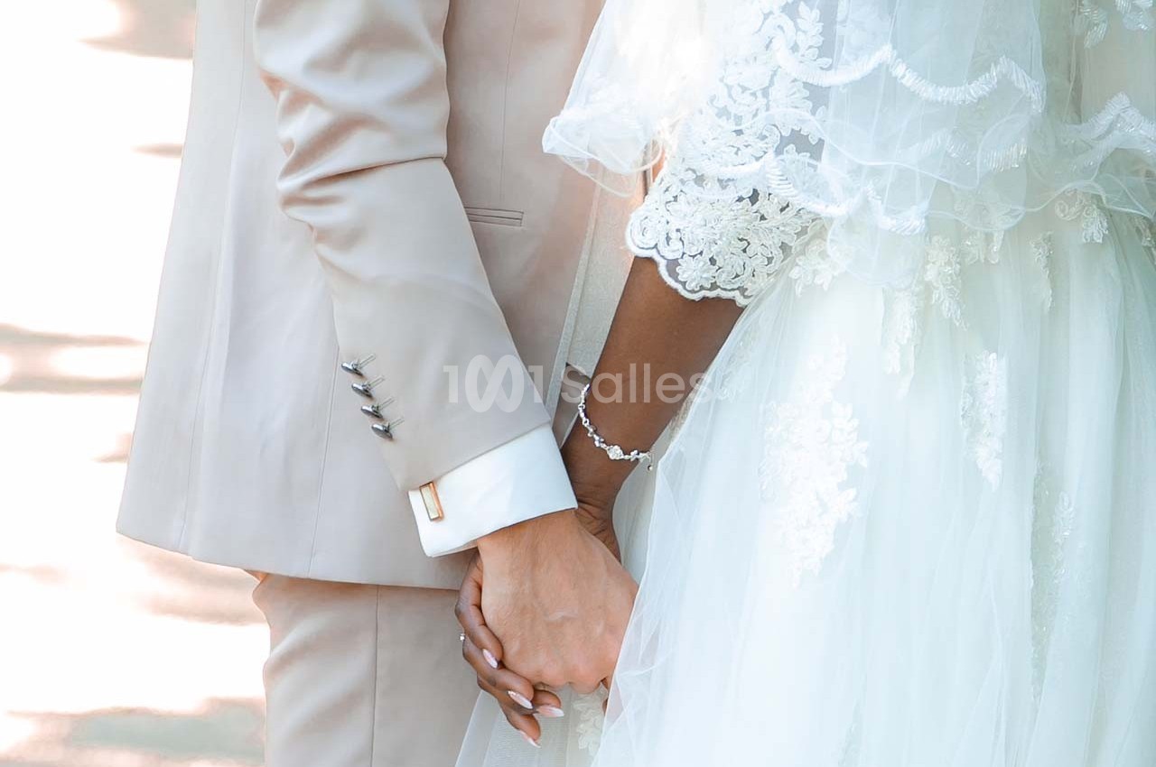 Un couple en tenue de mariage se tient la main, mettant en valeur des détails de la robe et du costume.