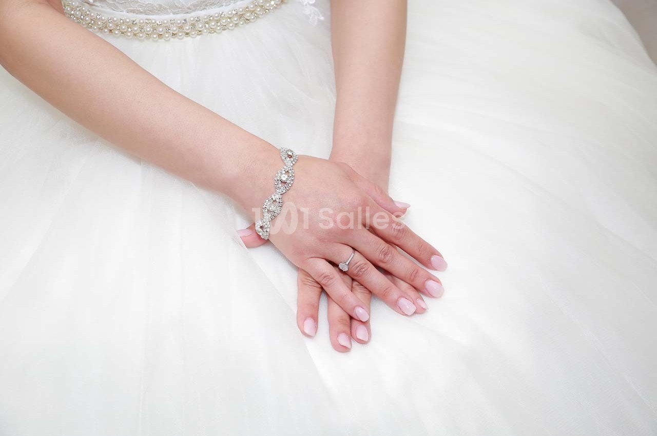 Mains croisées d'une femme portant une robe blanche, un bracelet argenté et une bague sur fond de tissu clair.