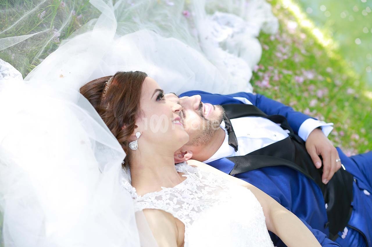 Un couple en tenue de mariage allongé sur l'herbe, entouré de pétales de fleurs et de voiles blancs.
