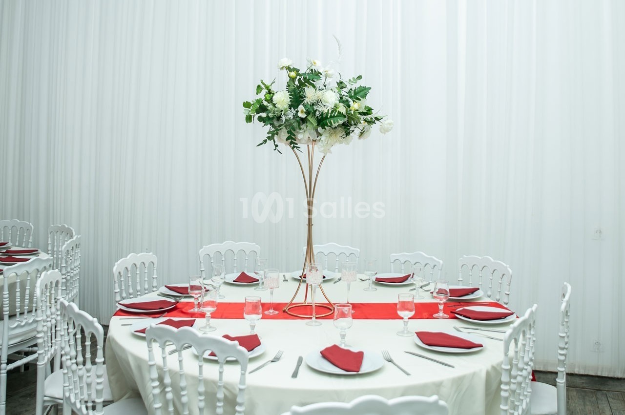 Table ronde décorée avec une nappe blanche, des serviettes rouges et un centre de table floral sur pied doré.