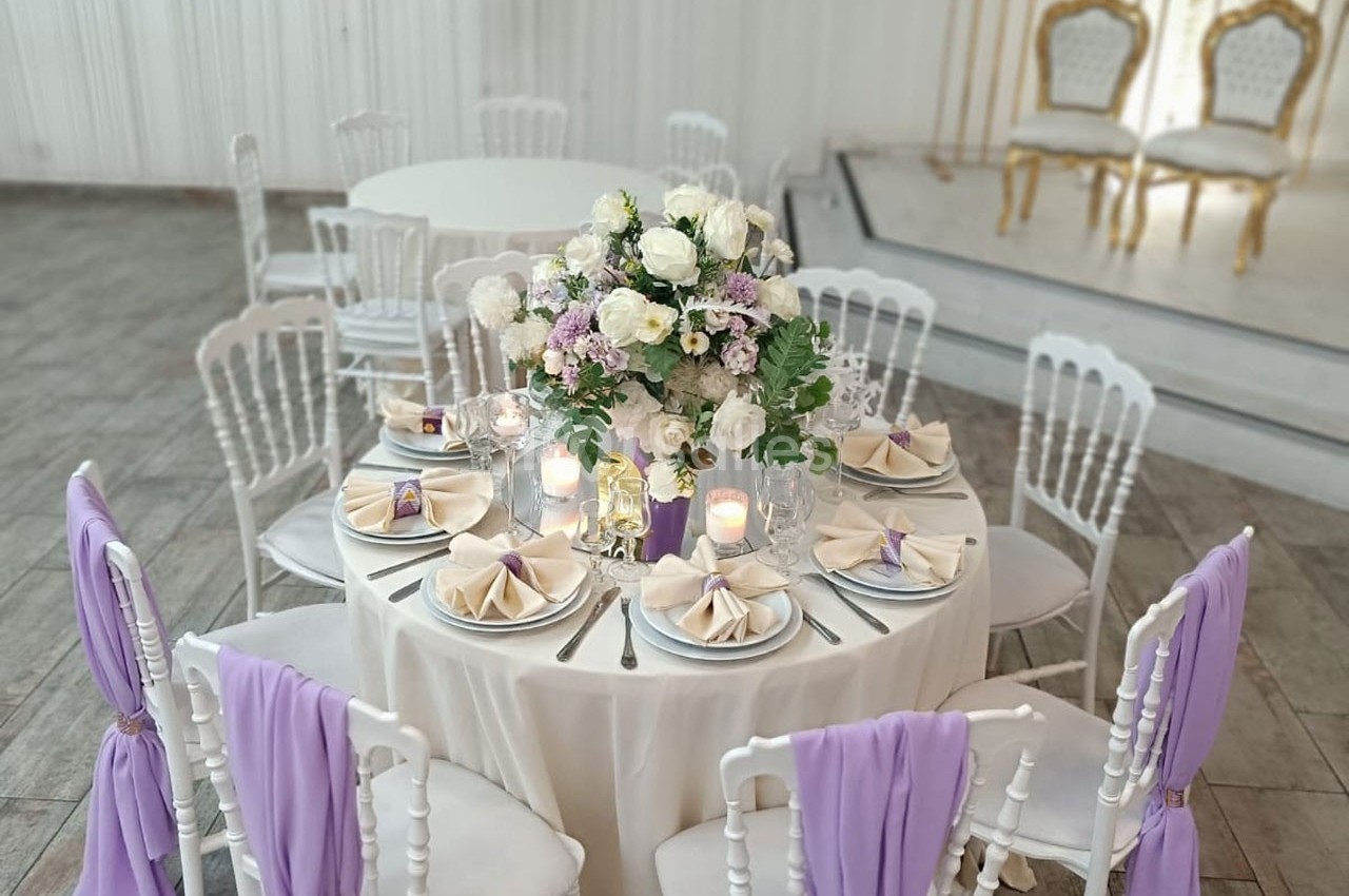 Table élégamment dressée avec des fleurs blanches et lilas, serviettes pliées et chaises décorées de tissu violet.