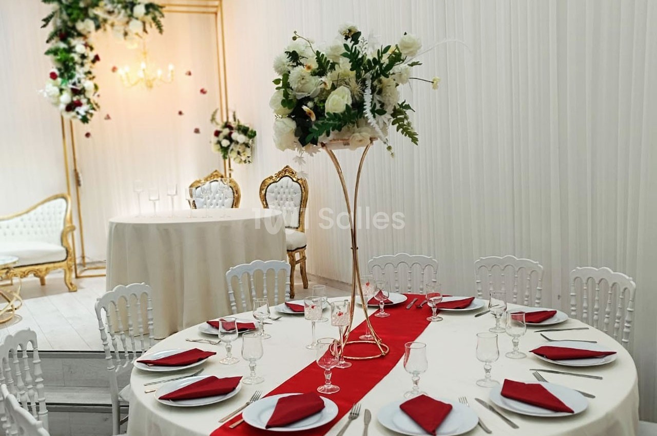 Salle de réception décorée avec des tables rondes, nappes blanches, chemins de table rouges et un centre floral élégant.