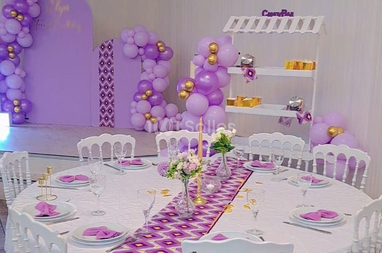 Table décorée avec nappe blanche, serviettes violettes, centre de table floral, et arrière-plan avec ballons violets et…