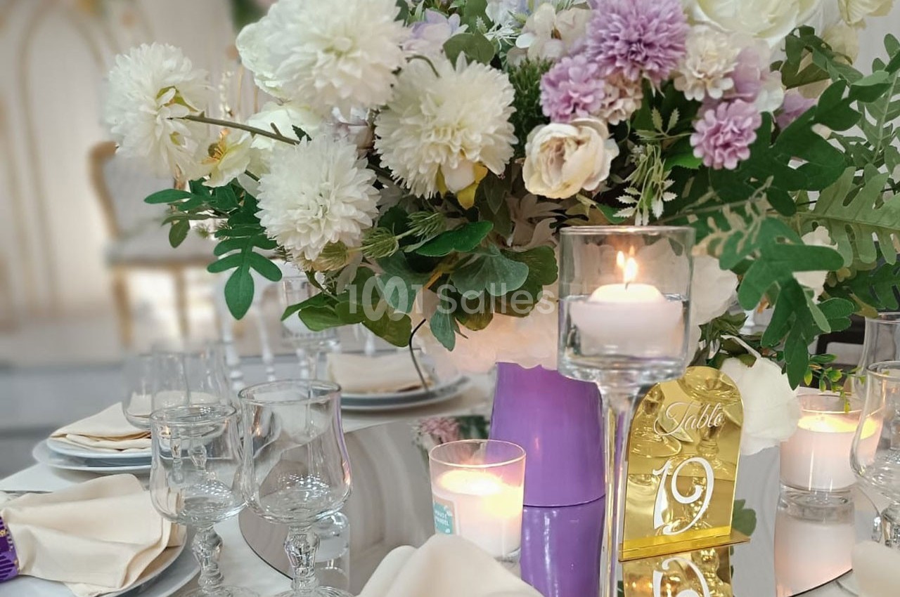 Centre de table floral avec des fleurs blanches et violettes, bougies allumées et numéro de table doré.