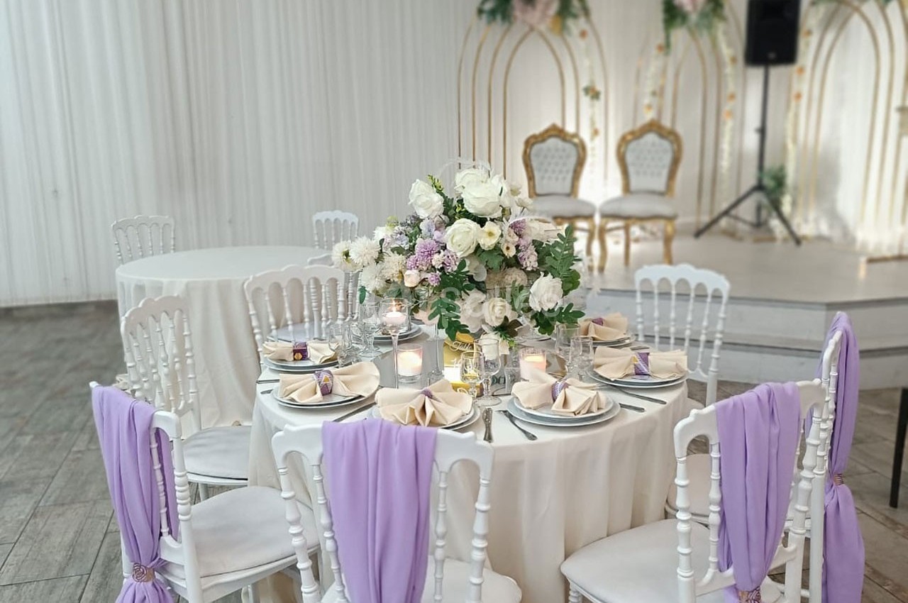 Table décorée pour un mariage avec nappes blanches, serviettes lilas, centre de table floral et chaises assorties.