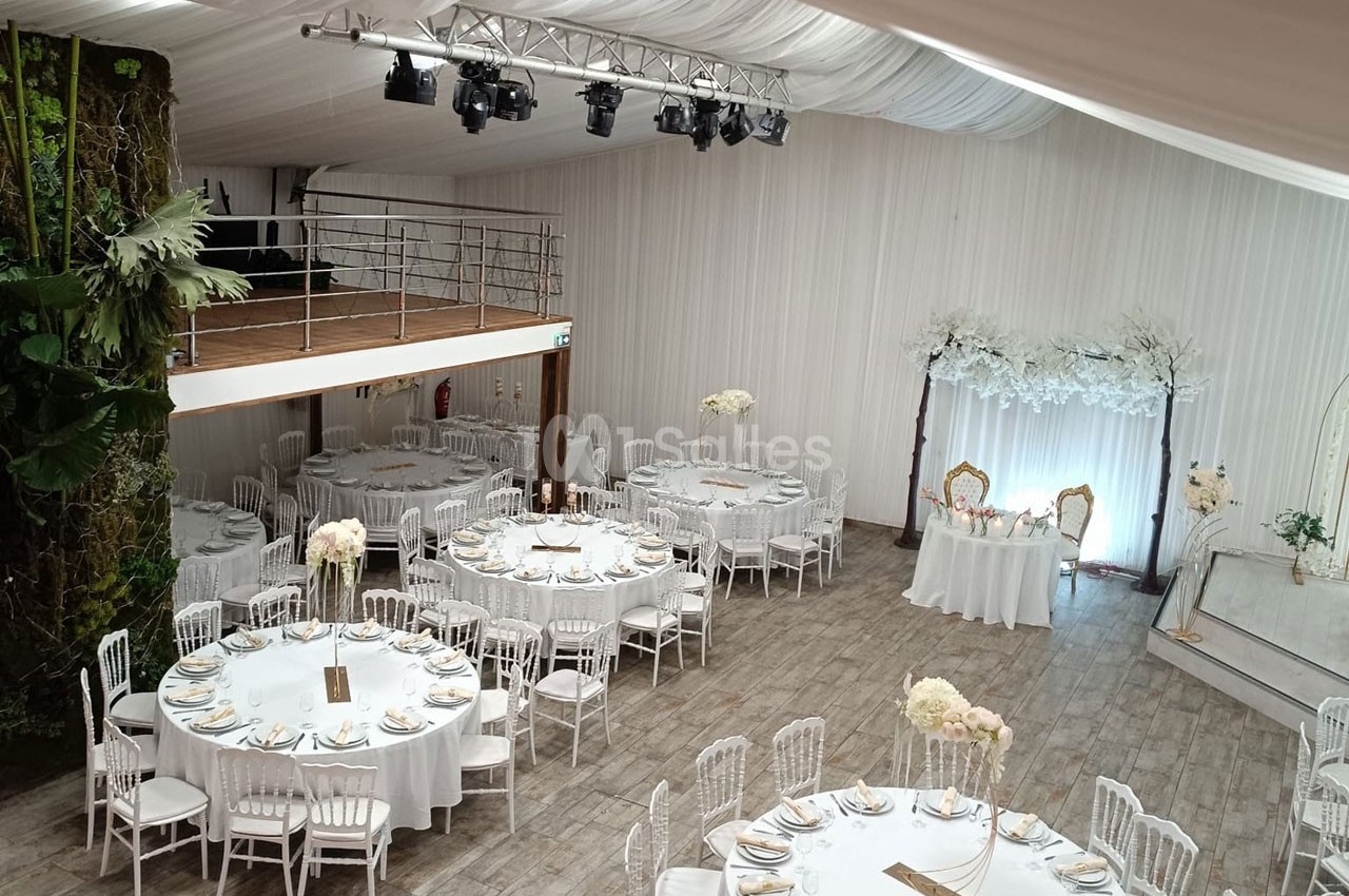 Salle de réception décorée avec des tables rondes dressées, des chaises blanches et une arche florale en arrière-plan.