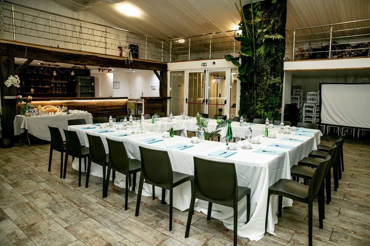 Salle aménagée pour une réunion avec des tables en U, nappes blanches, chaises noires et décor végétal en arrière-plan.