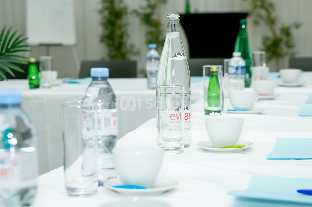 Bouteilles d'eau, tasses et documents disposés sur des tables blanches dans une salle de réunion lumineuse.