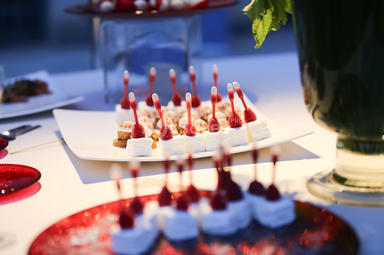 Assortiment de bouchées sucrées décorées de framboises et de pipettes, présenté sur des plateaux lors d'un événement.