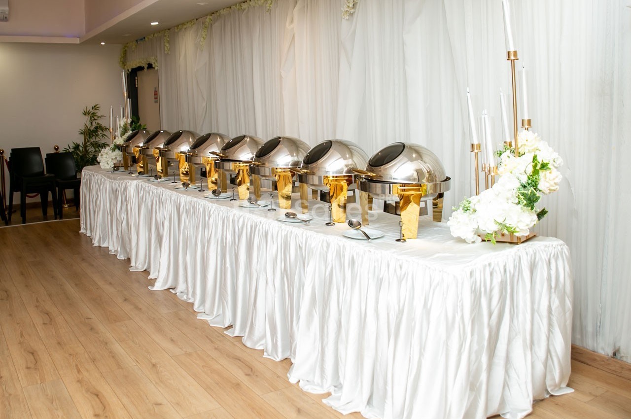Buffet avec plusieurs plats en chafing dishes alignés sur une table drapée de blanc, décorée de fleurs.