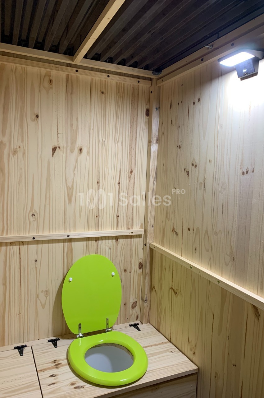 Toilettes sèches en bois clair avec abattant vert, éclairées par une lampe fixée au mur.