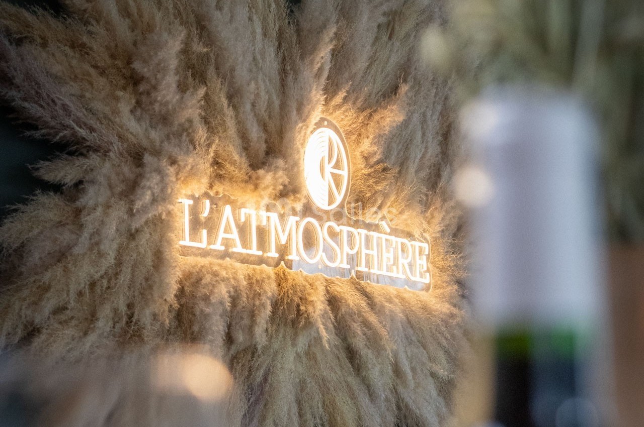 Enseigne lumineuse ’L'Atmosphère’ entourée de pampas beige sur un fond flou.