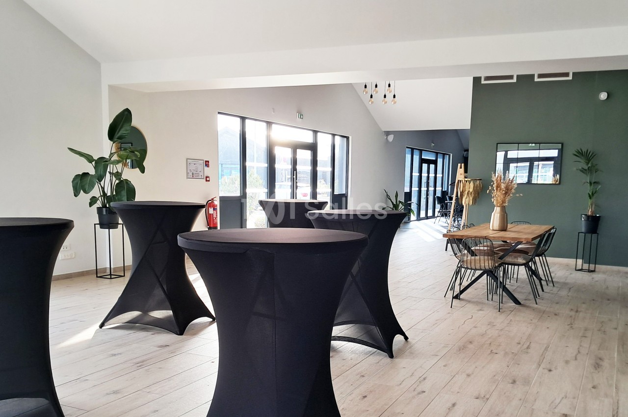 Salle lumineuse avec tables hautes noires, plantes décoratives et grande table en bois près de baies vitrées.