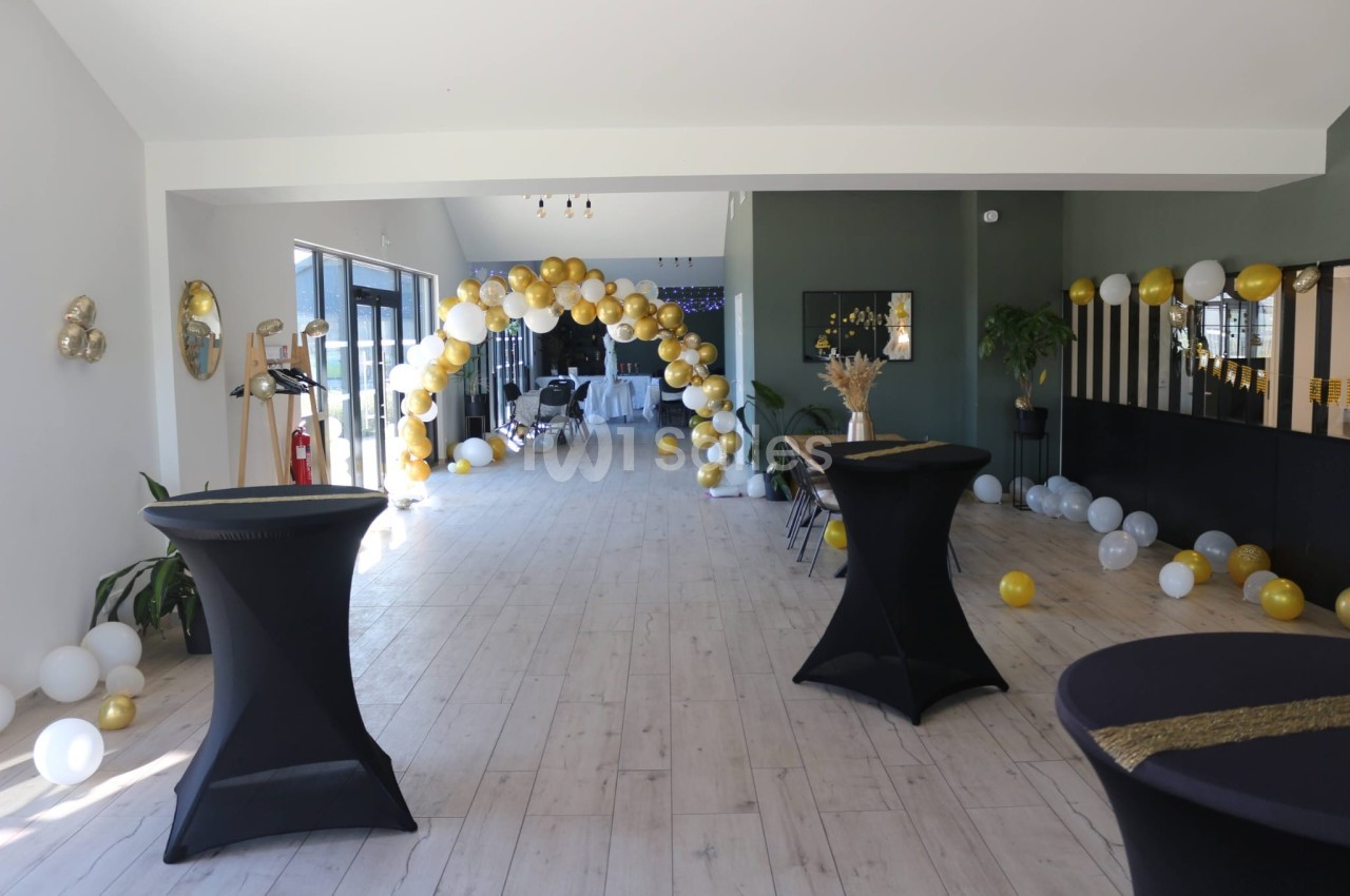 Salle décorée avec des ballons dorés et blancs, tables hautes noires et arche festive en arrière-plan.