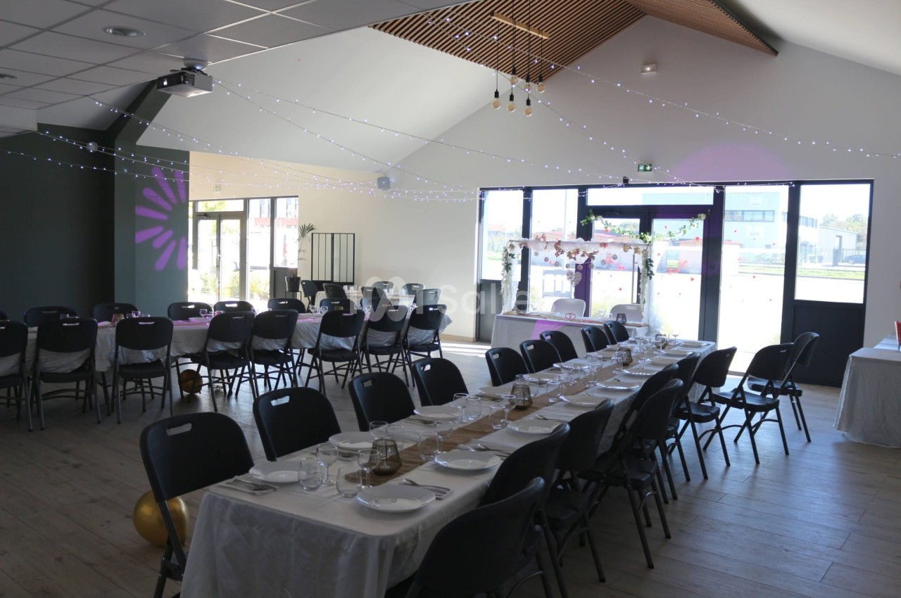 Salle de réception décorée avec des tables dressées, guirlandes lumineuses suspendues et lumière naturelle entrant par les…