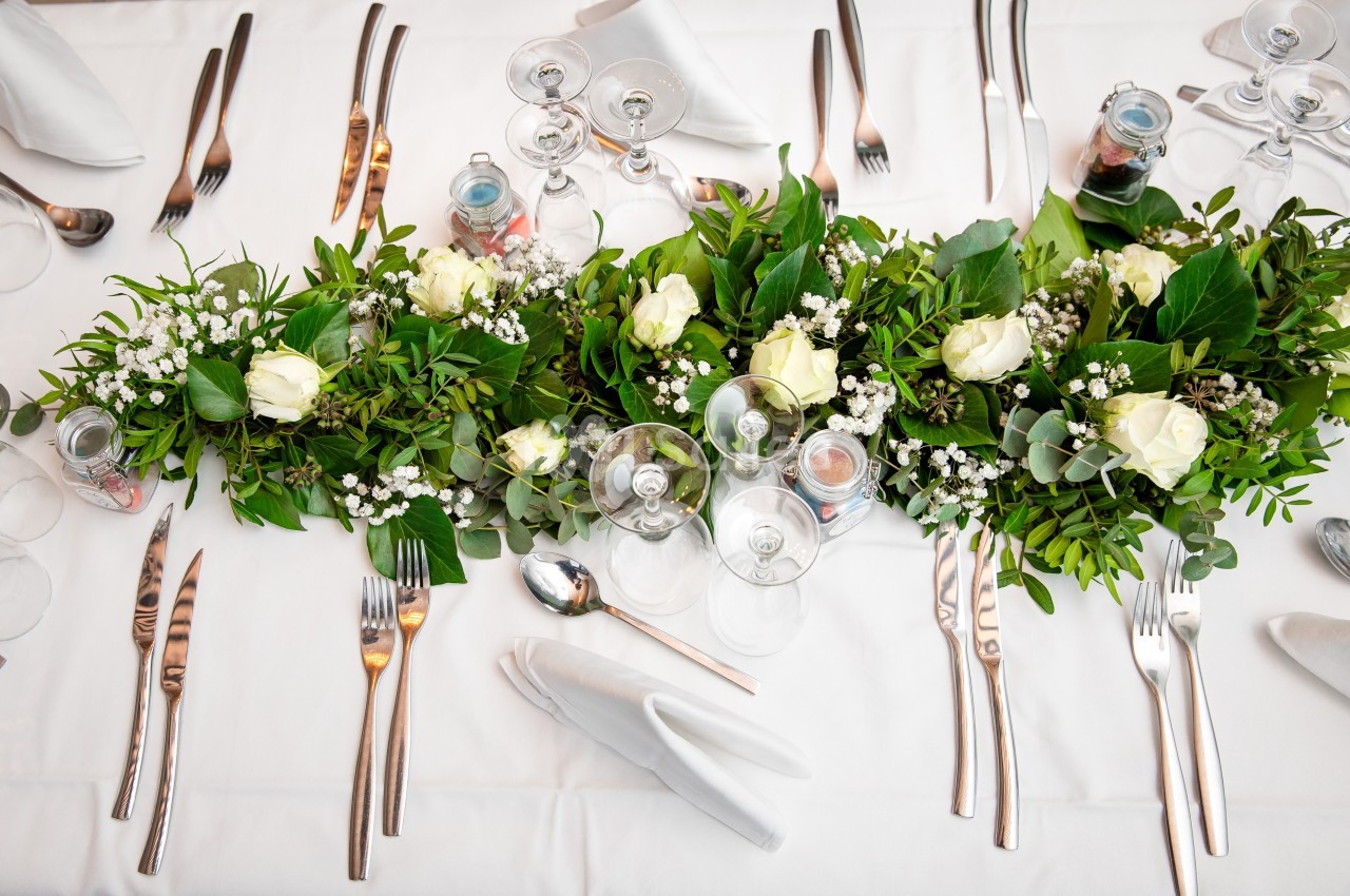 Centre de table avec des roses blanches, feuillage vert et vaisselle disposée sur une nappe blanche.