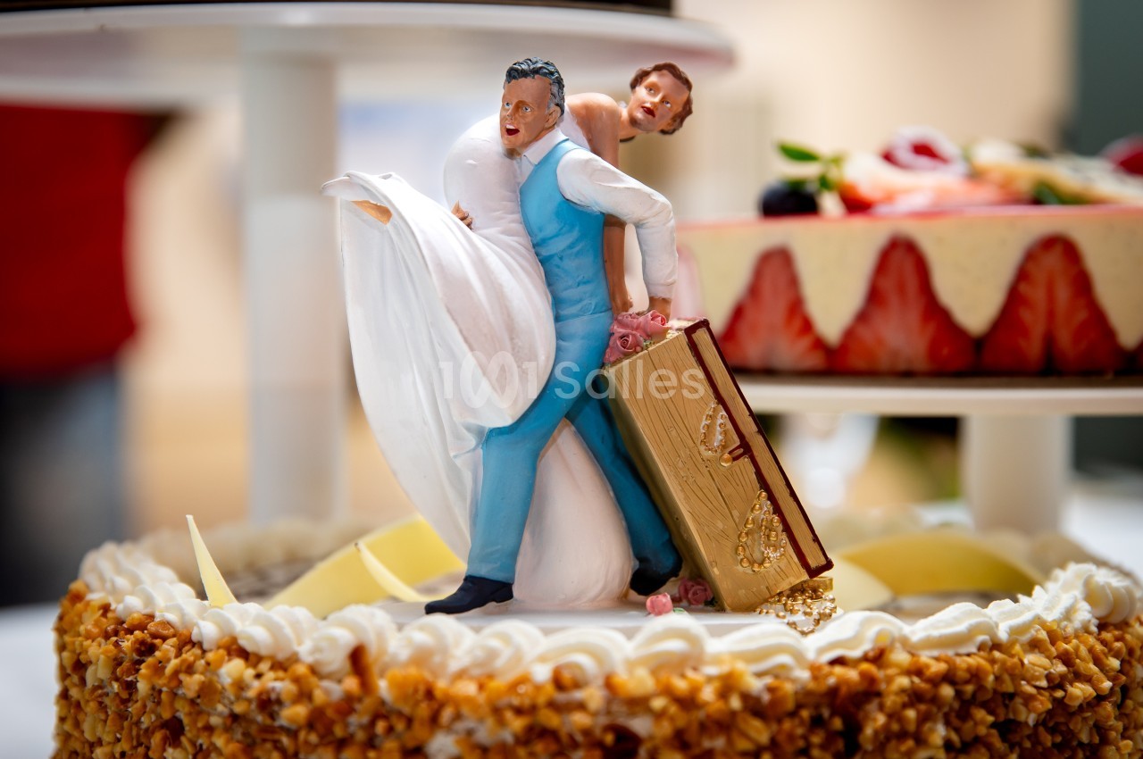 Figurines humoristiques de mariés sur un gâteau, le marié portant une valise et la mariée accrochée à lui.