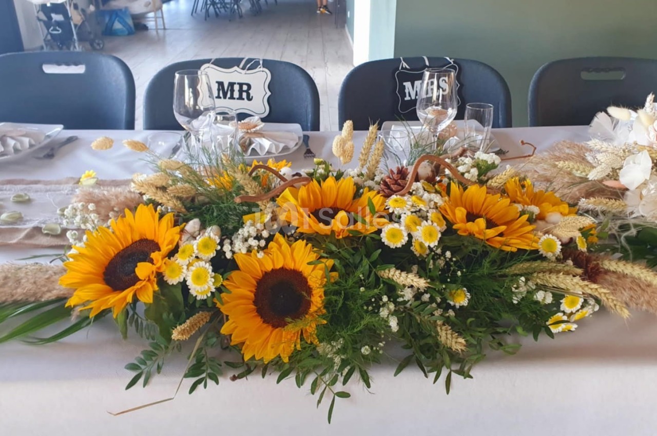 Centre de table avec des tournesols, des marguerites et des herbes séchées, disposé sur une table de réception.