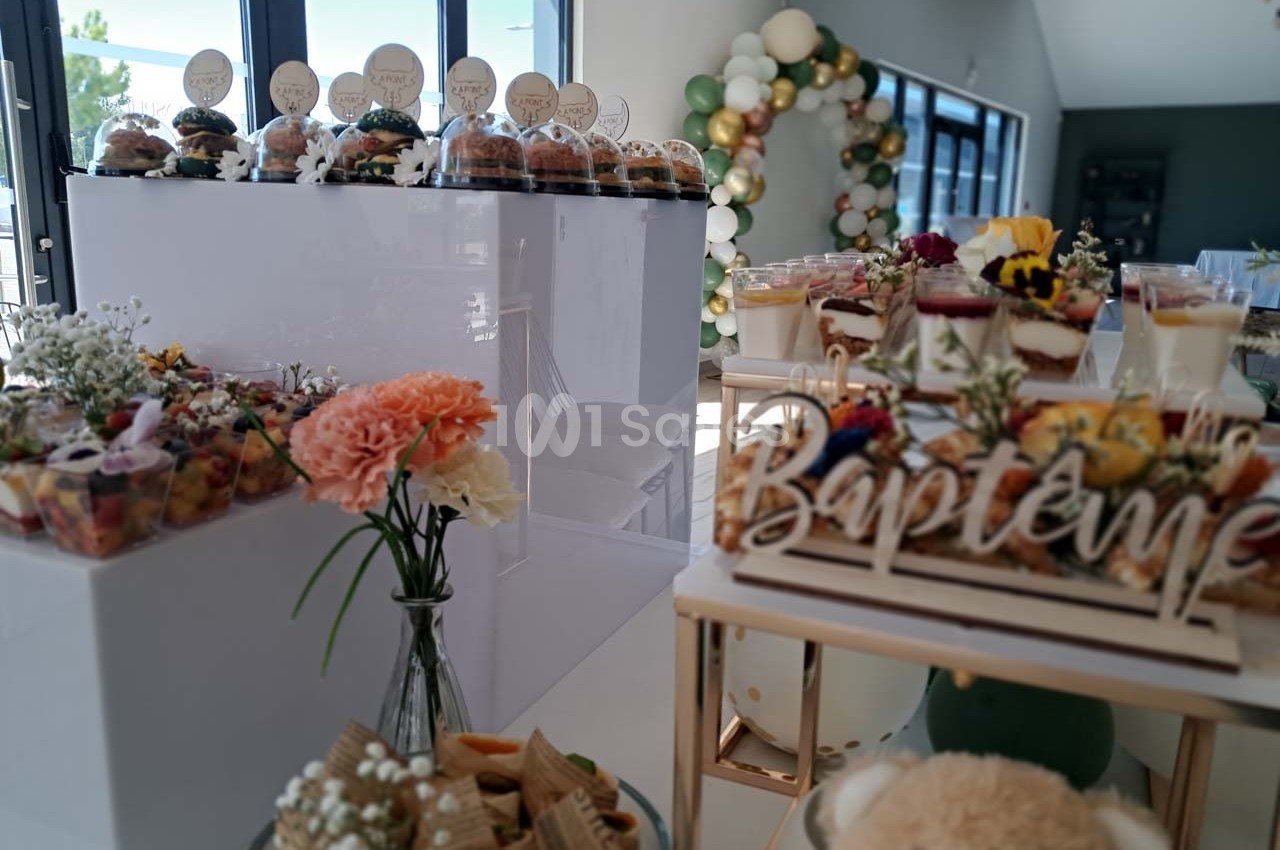 Buffet de baptême avec desserts variés, fleurs décoratives et ballons en arrière-plan dans une salle lumineuse.