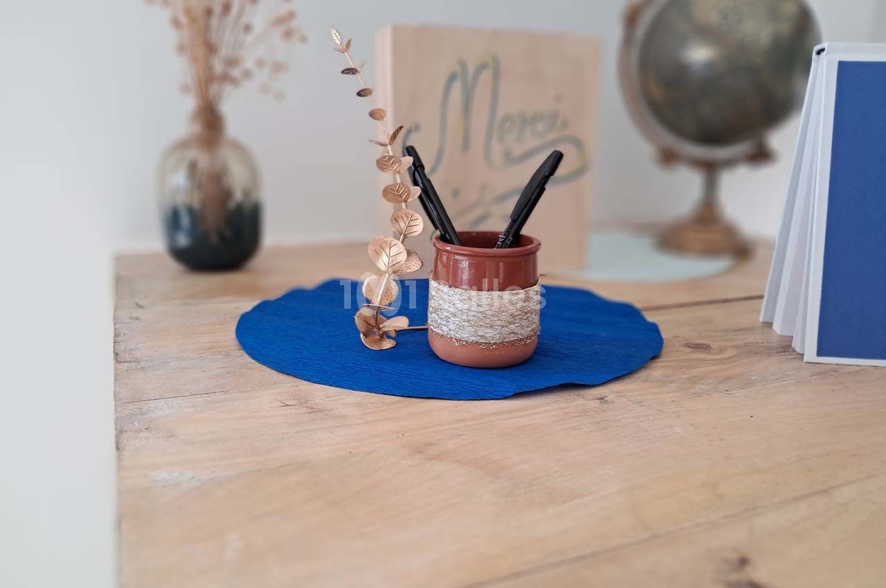 Pot en céramique contenant des stylos et une branche décorative, posé sur un napperon bleu sur une table en bois.