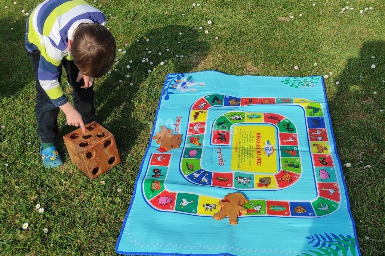 Un enfant joue sur une pelouse avec un dé en bois et un tapis de jeu coloré représentant un parcours.