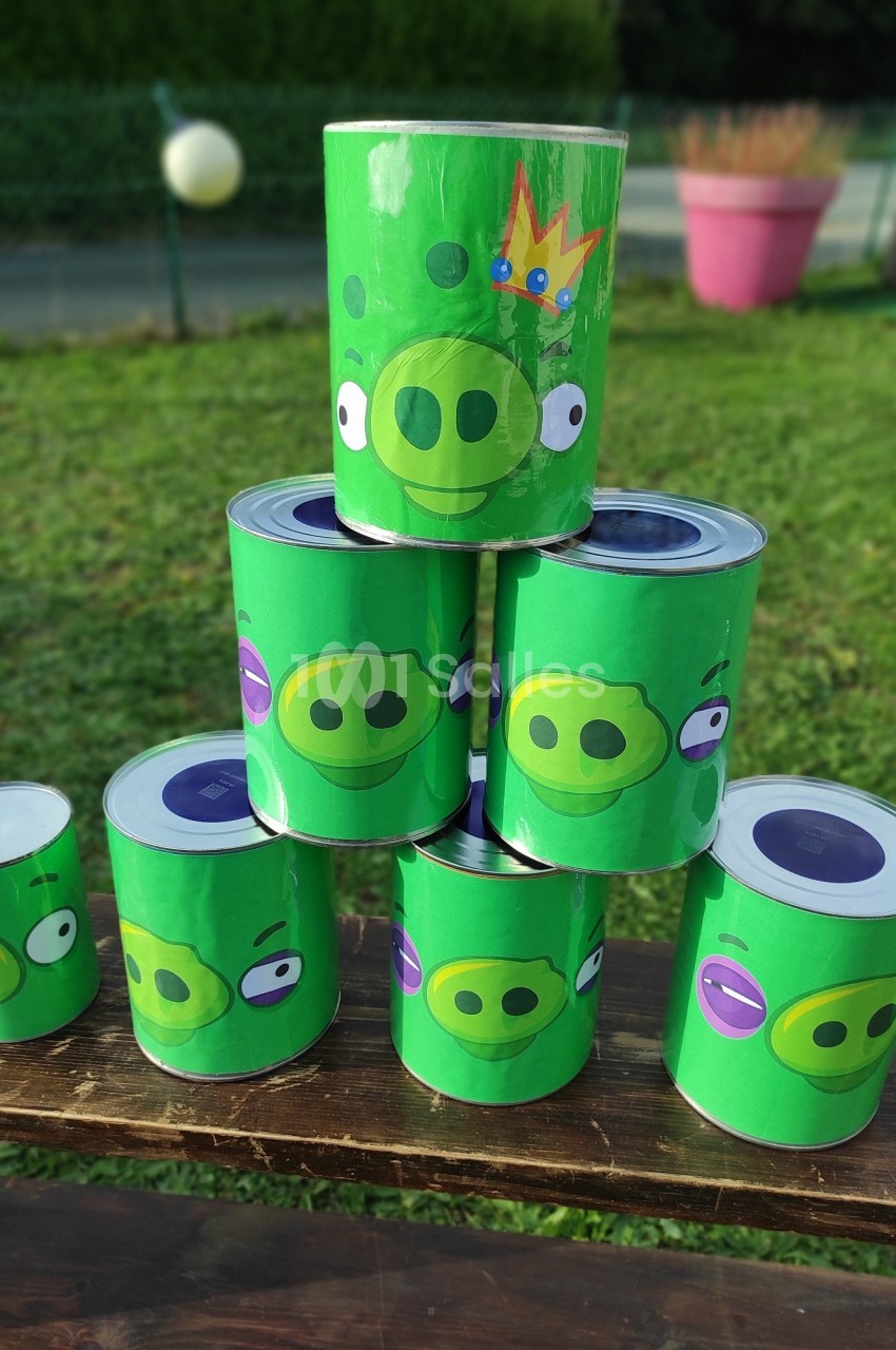 Six boîtes de conserve décorées de visages de cochons verts empilées en pyramide sur une table en extérieur.
