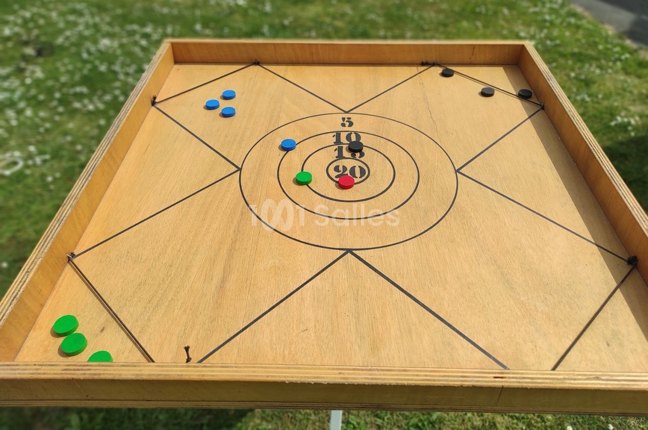 Plateau de carrom en bois avec des pions colorés dispersés, posé sur une pelouse verte.