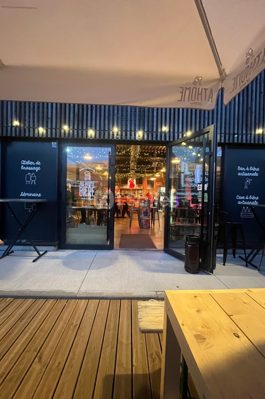 Entrée d'un bar avec terrasse en bois, portes vitrées ouvertes et intérieur éclairé par des guirlandes lumineuses.