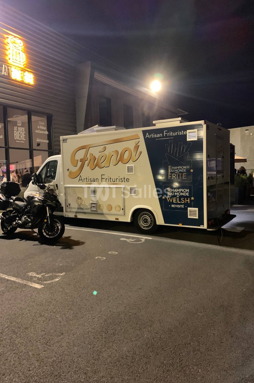 Camion de friterie artisanale stationné de nuit devant un bâtiment éclairé, avec une moto garée à côté.