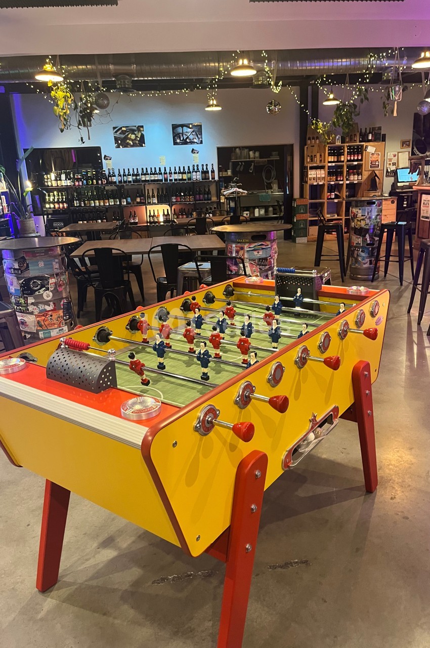 Baby-foot jaune dans un bar avec tables hautes, étagères de bouteilles et éclairage tamisé en arrière-plan.