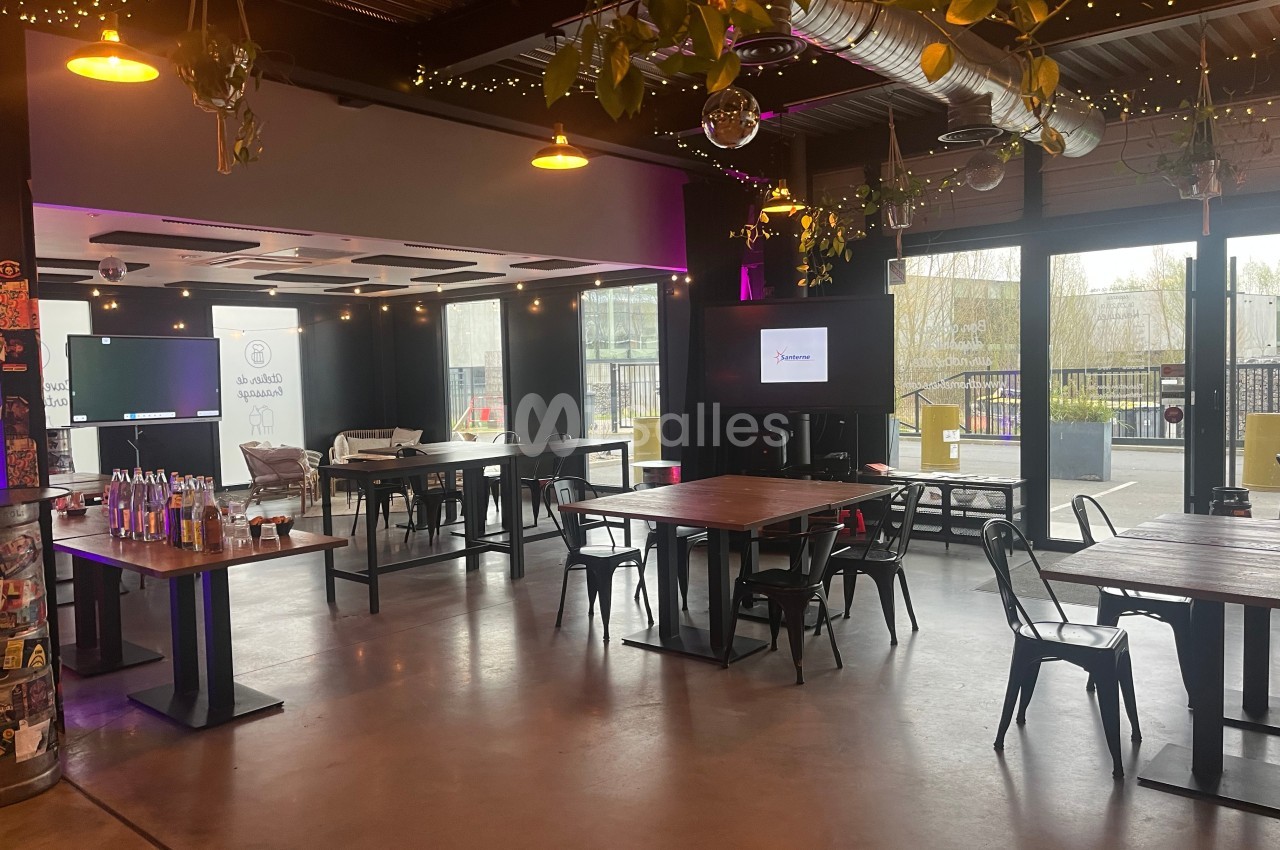 Salle lumineuse avec tables en bois, chaises métalliques, décorations suspendues et écrans allumés en arrière-plan.