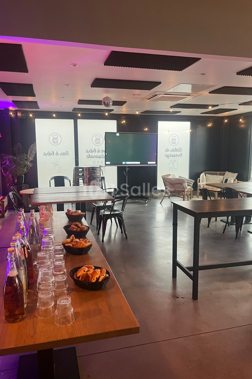 Salle lumineuse aménagée pour un événement, avec tables, chaises, écran et buffet de boissons et viennoiseries.