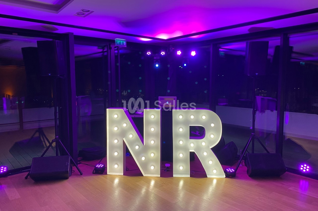Lettres lumineuses ’NR’ devant une baie vitrée, entourées de projecteurs et éclairées par une lumière violette.