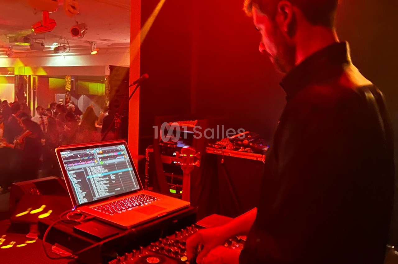 Un DJ mixe de la musique sur un ordinateur portable et une table de mixage dans une salle éclairée de lumières rouges.