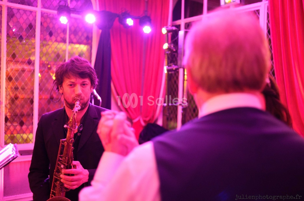 Un musicien joue du saxophone dans une salle éclairée de lumières roses, face à une personne de dos.