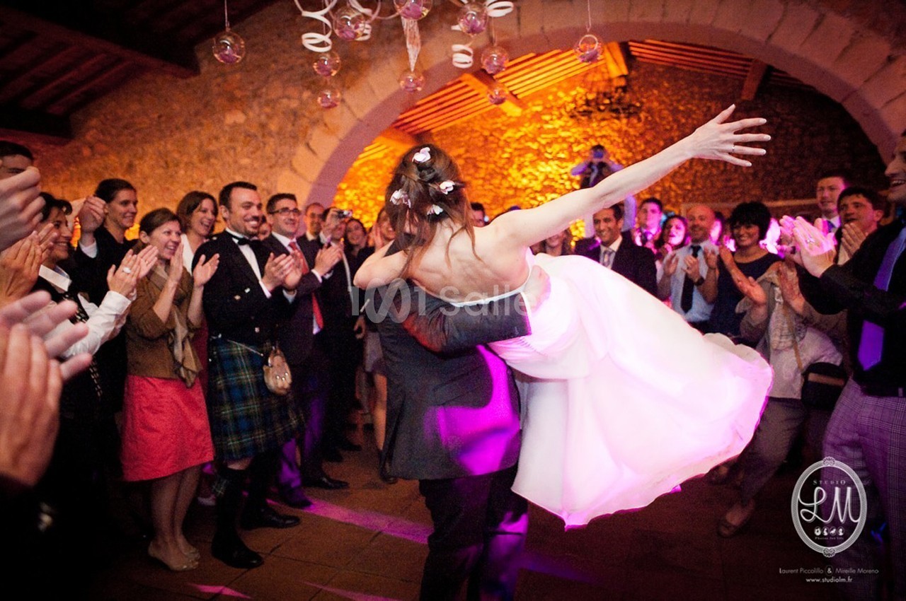 Un couple de mariés danse devant des invités applaudissant dans une salle en pierre éclairée de lumières chaudes.