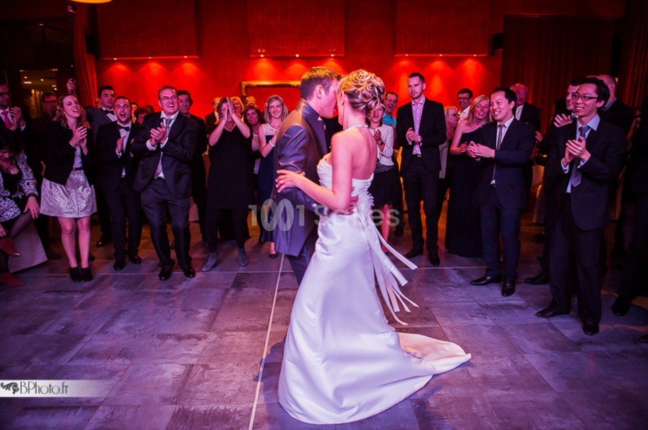 Un couple danse au centre d'une salle, entouré d'invités applaudissant dans une ambiance festive et éclairage chaleureux.