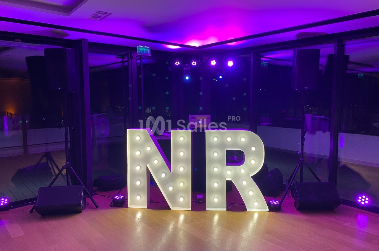 Lettres lumineuses ’NR’ devant une baie vitrée, entourées de projecteurs et éclairées par une lumière violette.