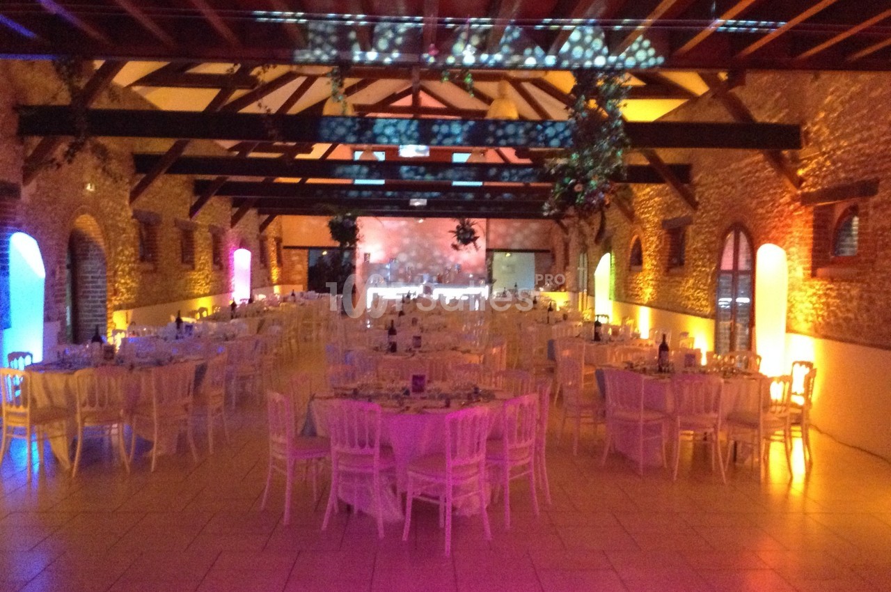 Salle de réception en pierre avec poutres apparentes, tables rondes dressées et éclairage coloré.