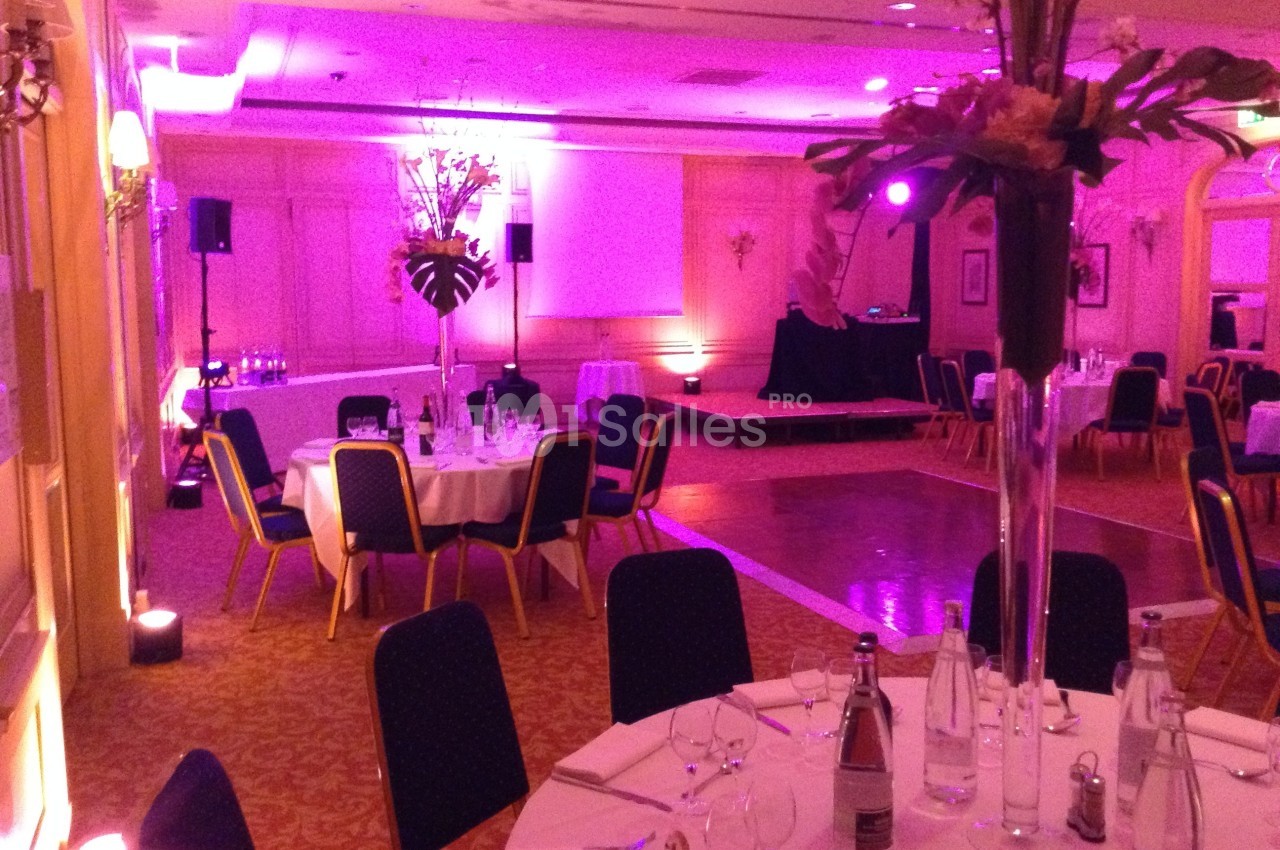 Salle de réception avec tables rondes dressées, éclairage tamisé rose et espace pour une scène.