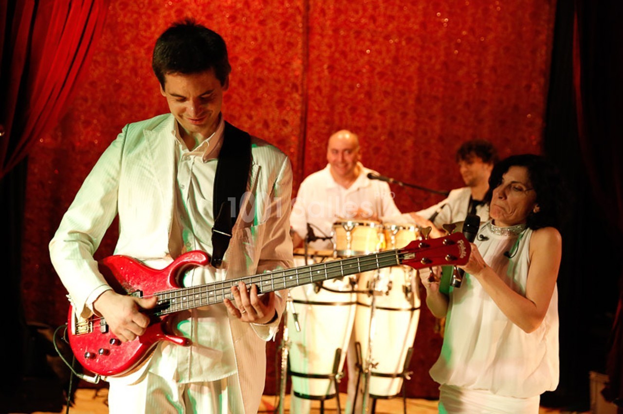Un musicien joue de la guitare électrique rouge sur scène, accompagné d'un groupe en arrière-plan.