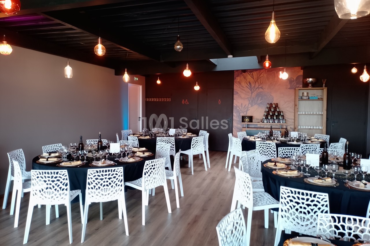 Salle de réception moderne avec tables dressées, chaises blanches ajourées et éclairage suspendu.