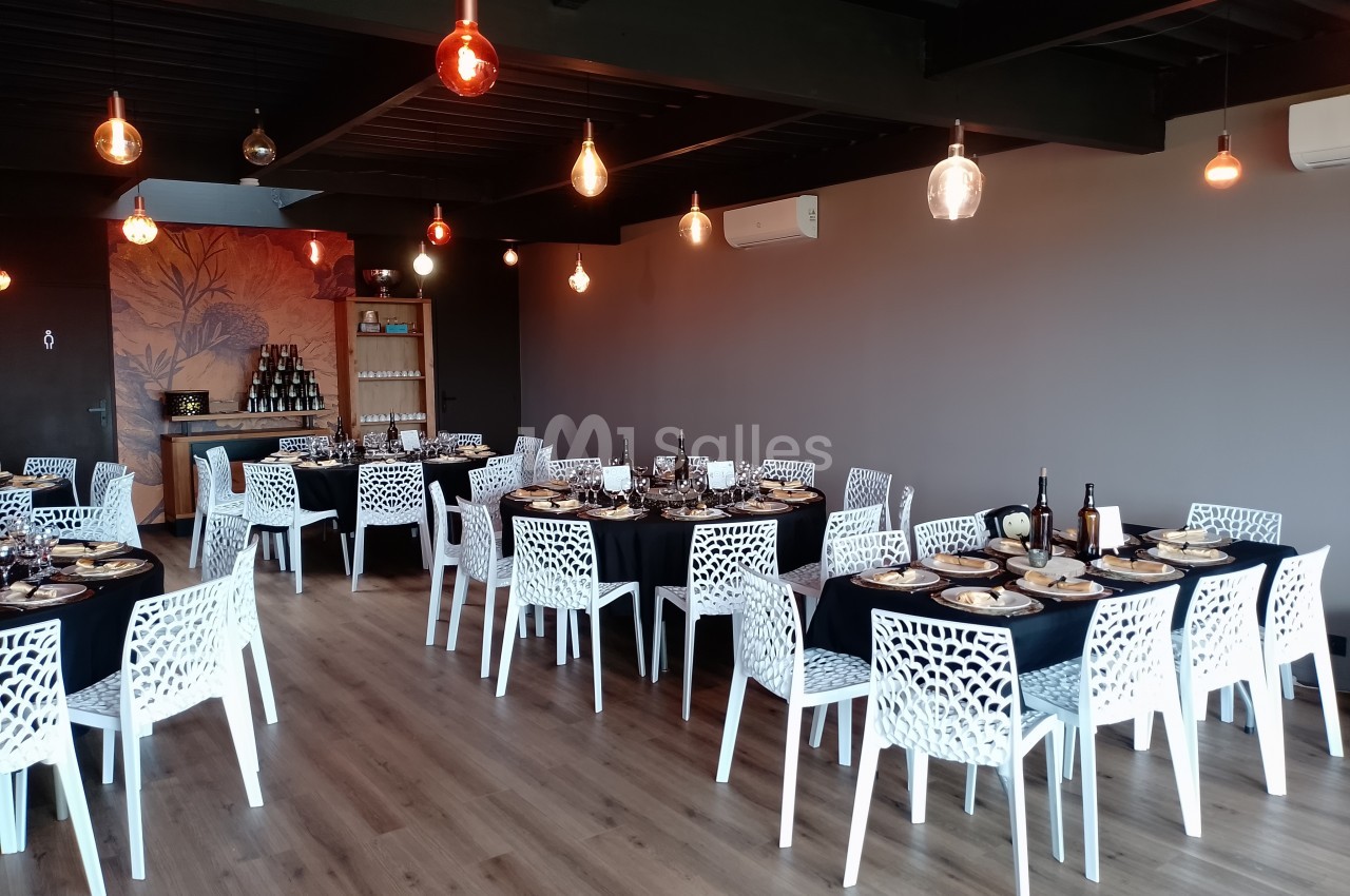 Salle de réception avec tables dressées, chaises blanches ajourées et éclairage suspendu dans un décor moderne.