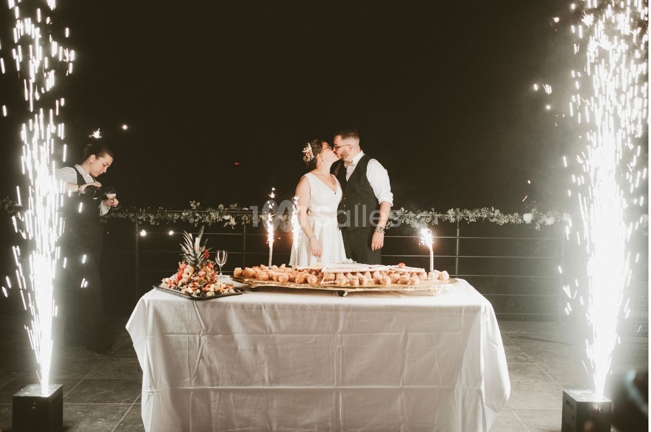 Un couple de mariés se tient près d'une table garnie de desserts, entouré de fontaines lumineuses.