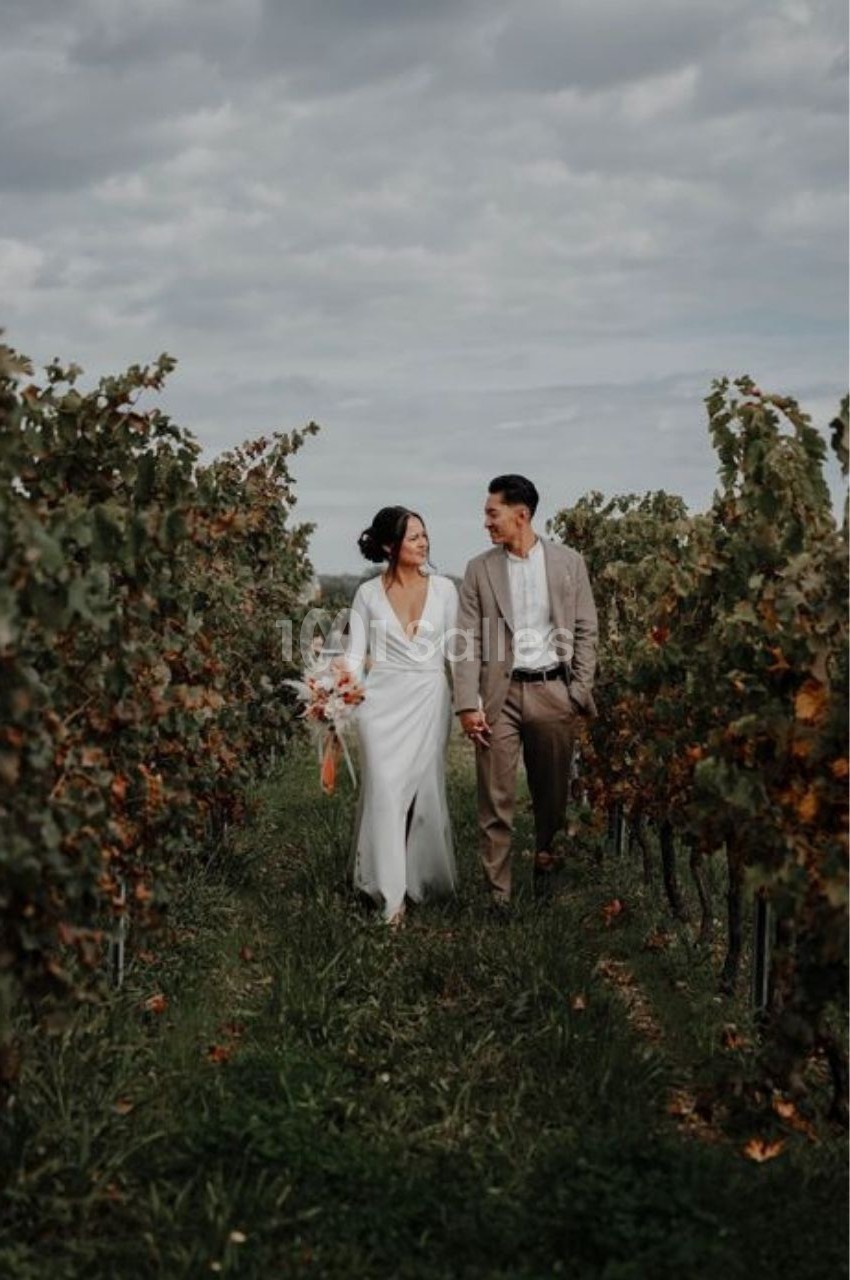 Un couple marche main dans la main dans un vignoble sous un ciel nuageux.