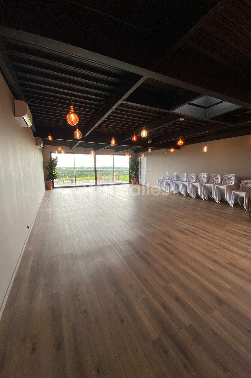 Salle spacieuse avec parquet, chaises alignées à droite, grandes baies vitrées donnant sur un extérieur verdoyant.