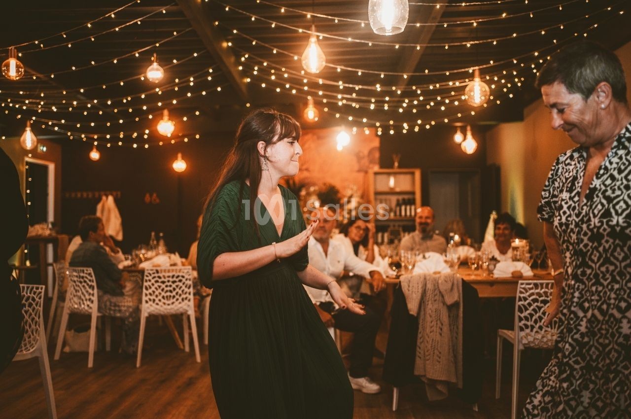 Une femme danse dans une salle décorée de guirlandes lumineuses, entourée de convives assis à des tables.