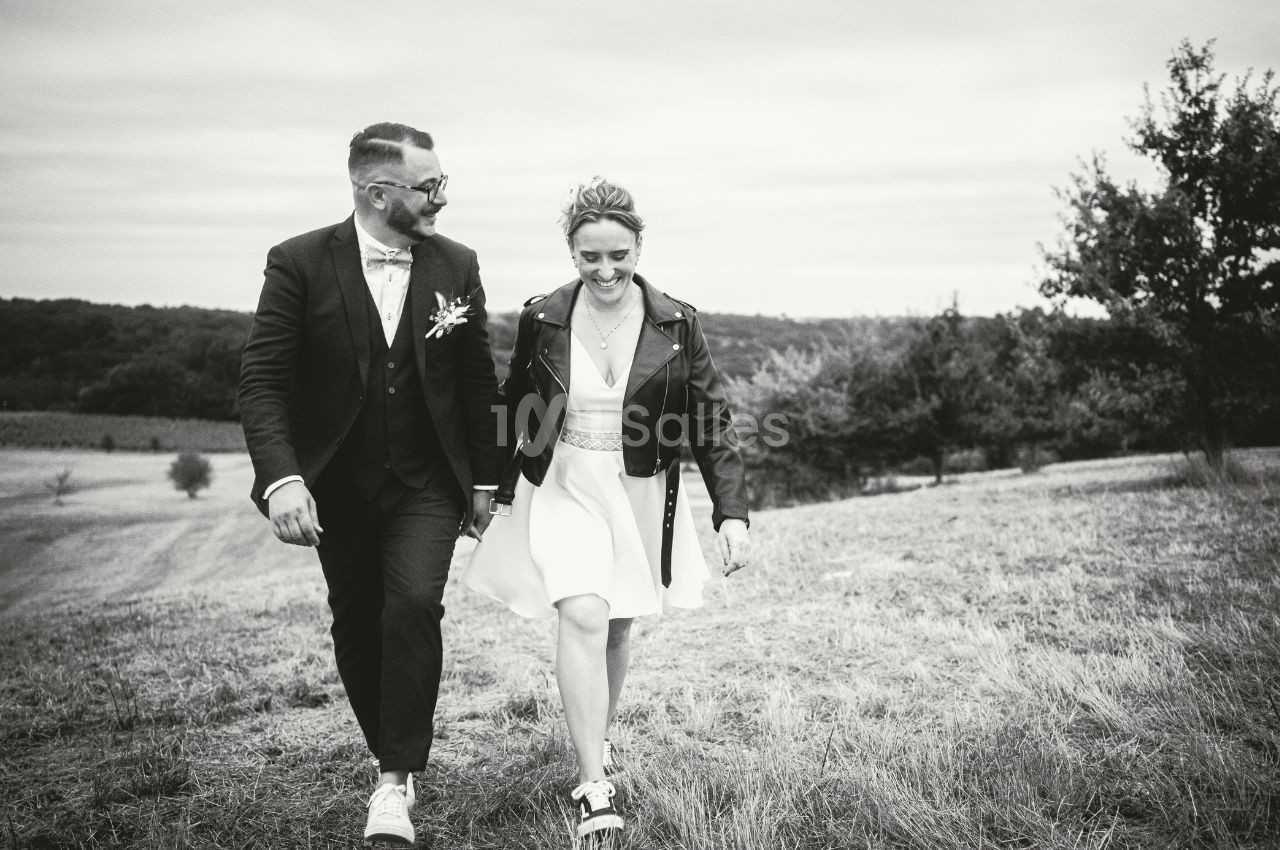Un couple souriant marche dans un champ, elle en robe et blouson, lui en costume, sous un ciel nuageux.