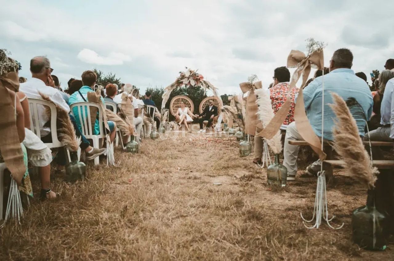Cérémonie de mariage en plein air avec des invités assis et une arche décorée au centre sous un ciel nuageux.